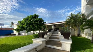 A building with a white exterior, a covered outdoor seating area, and a paved pathway leading to the entrance. - 1415 2-bedroom apartment in serene Manzanillo with AC for comfort (Manzanillo)