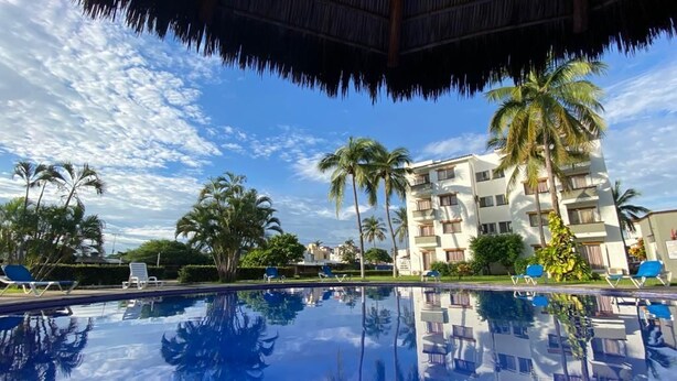 A swimming pool with clear reflections, palm trees, and a building with balconies. - 1415 2-bedroom apartment in serene Manzanillo with AC for comfort (Manzanillo)