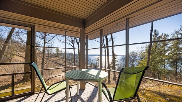 Un espacio al aire libre cubierto con una mesa y sillas, ventanas amplias y vistas a árboles y un cuerpo de agua.