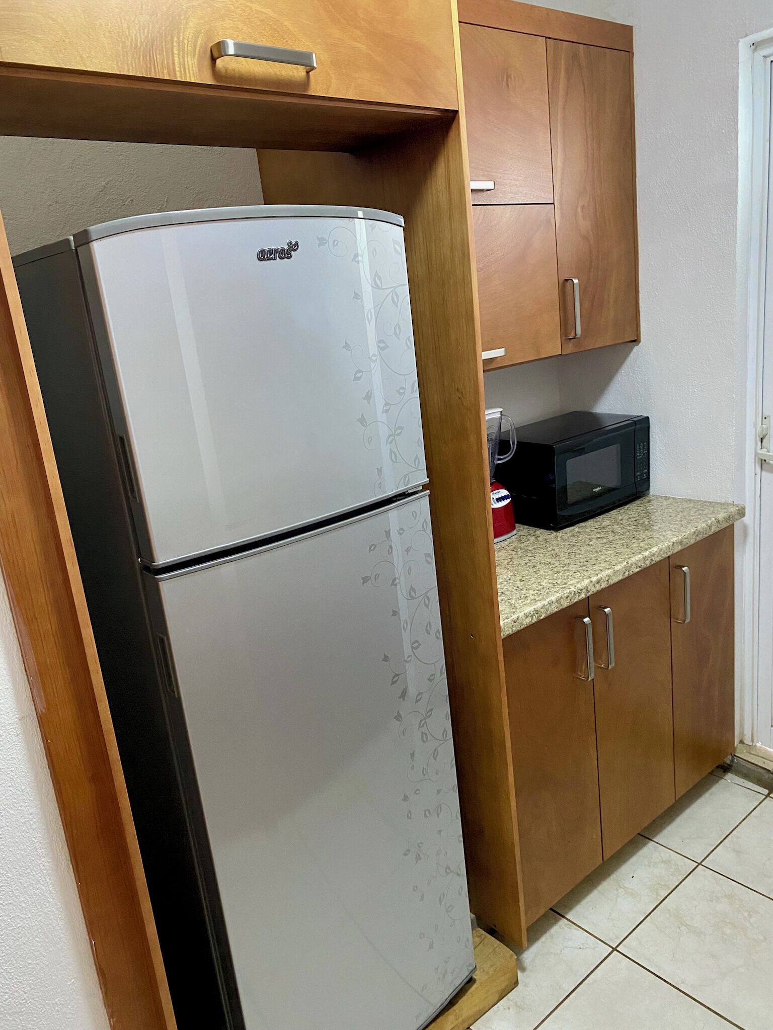 Eine Küche mit Kühlschrank, Mikrowelle, Holzschränken und Fliesenboden.