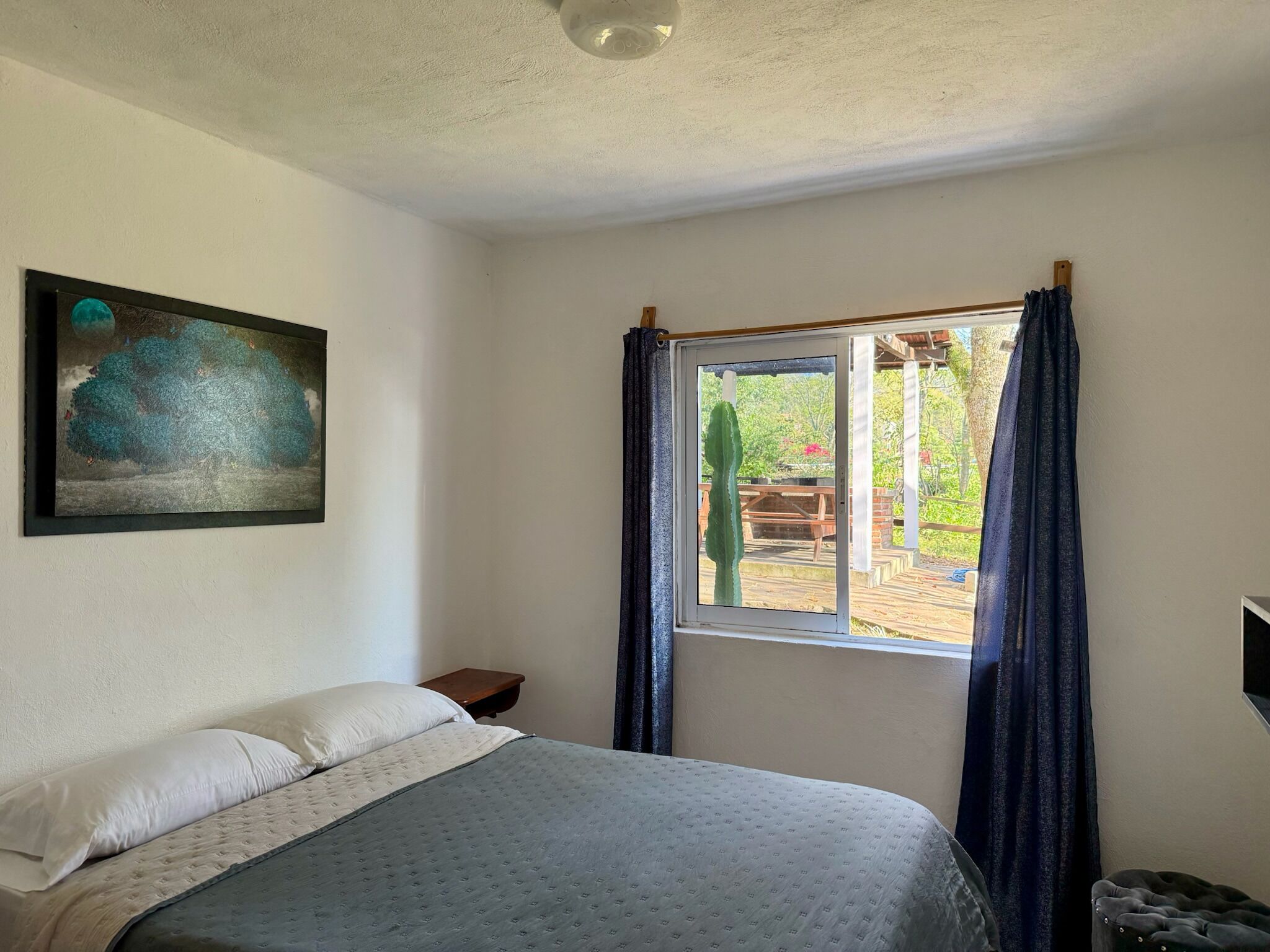 Un dormitorio con cama, una ventana con cortinas, una mesita pequeña y un cuadro en la pared.