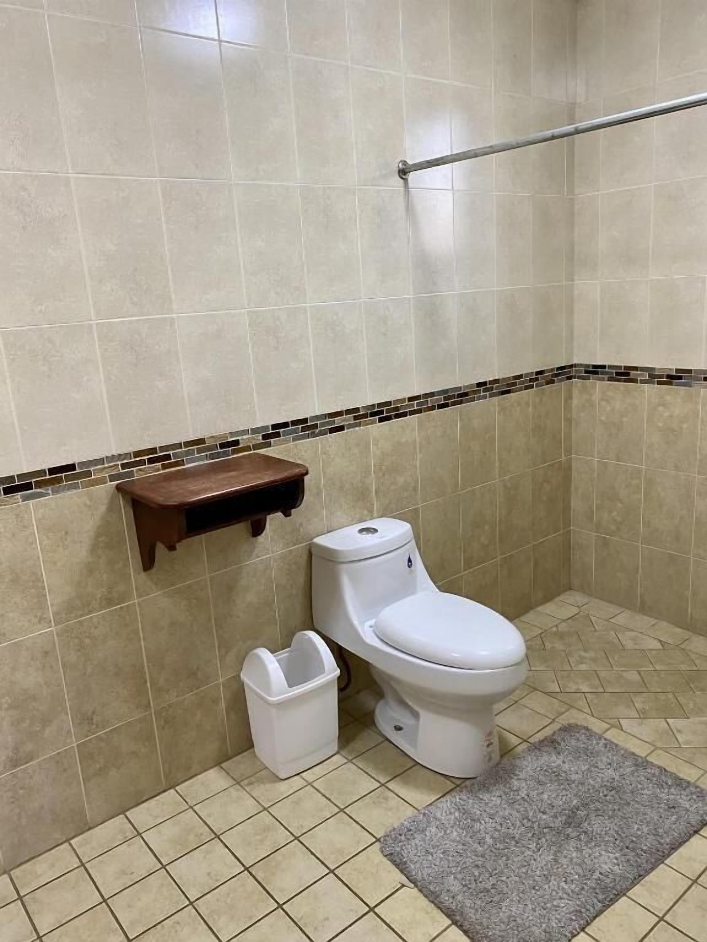 Un baño con inodoro, un cubo de basura, un pequeño estante de madera y una barra de apoyo.