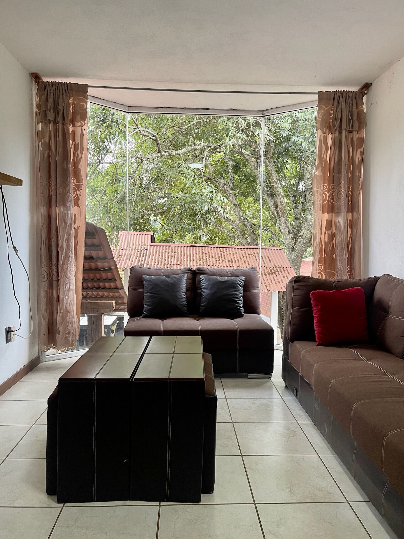 Un salon comprenant un canapé, un pouf et une table basse, de grandes fenêtres avec des rideaux, et une vue sur un bâtiment aux toits rouges à l’extérieur.
