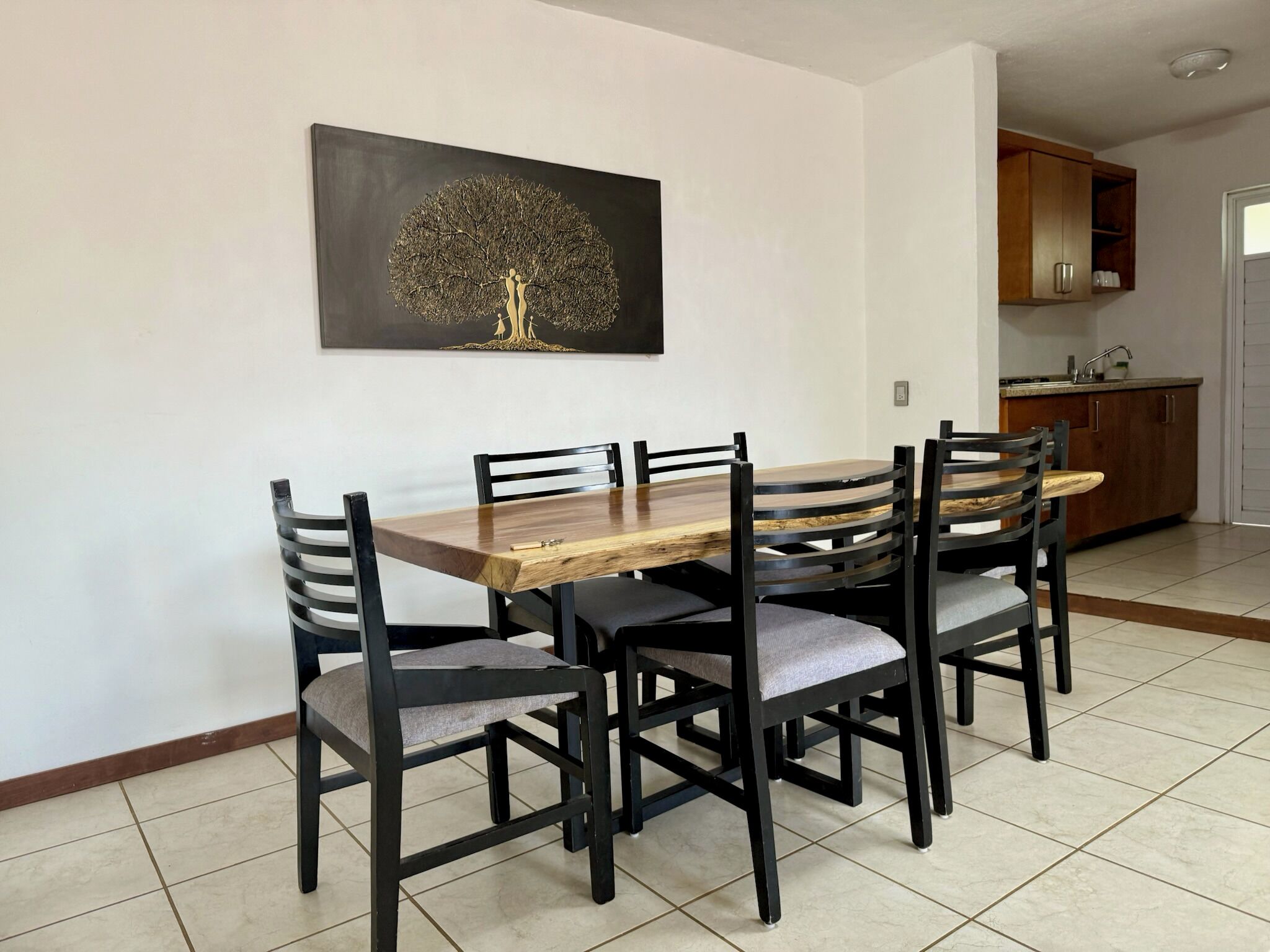 Uma área de refeições com mesa e cadeiras de madeira, cozinha ao fundo e uma obra de arte na parede.