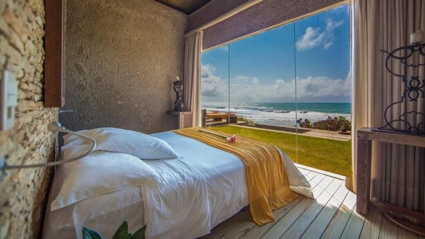 Une chambre avec une grande fenêtre donnant sur l’océan, un lit avec du linge de maison blanc et un plafond en bois.