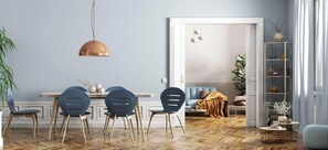 Un comedor con piso de madera en diseño espiga, una lámpara colgante de cobre y una puerta blanca que da a una sala de estar con sofá y planta.