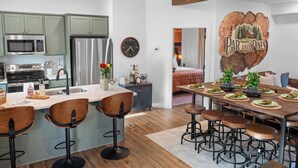 Cocina moderna con armarios verdes, refrigerador de acero inoxidable y una zona de comedor con mesa de madera y taburetes de bar.