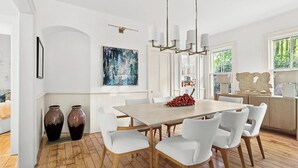 Een eetruimte met een houten tafel, witte stoelen en een kroonluchter. Op de vloer staan twee grote vazen, aan de muur hangt een schilderij en er is een raam met uitzicht op groen.