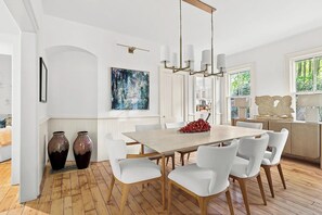 A dining area with a wooden table, white chairs, and a chandelier. There are two large vases on the floor, a painting on the wall, and a window with a view of greenery. - Peaceful 4-bedroom house with AC in charming Bridgehampton (Bridgehampton)
