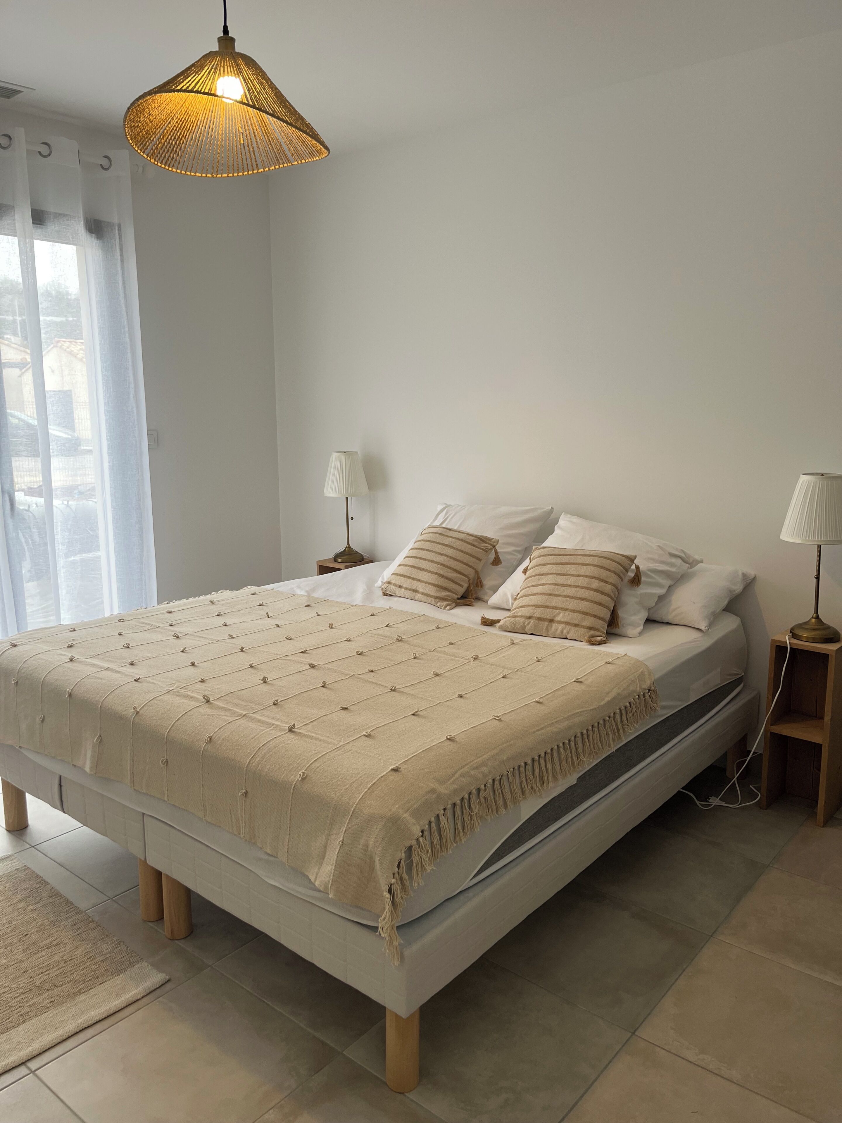 Una cama bien tendida con una manta y almohadas color beige, una lámpara colgante y una mesita de noche con lámpara.