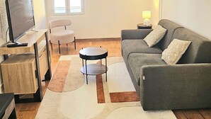 A living room with a grey sofa, a small round table, a TV on a wooden stand, a window with blinds, and a lamp on a side table. - Cosy T2 in downtown Le Mans, 15 minutes from the 24h circuit (Le Mans)