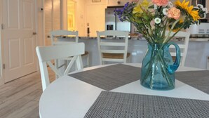 Un comedor con una mesa, sillas, un jarrón de flores y una cocina al fondo.