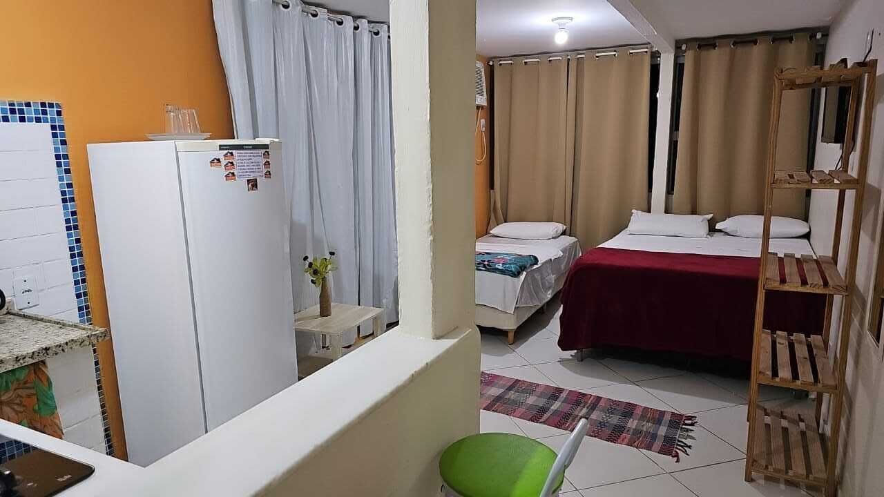 Une chambre d’hôtel compacte comprenant un coin cuisine, un lit recouvert d’une courtepointe rouge, une étagère en bois et une petite chaise verte.