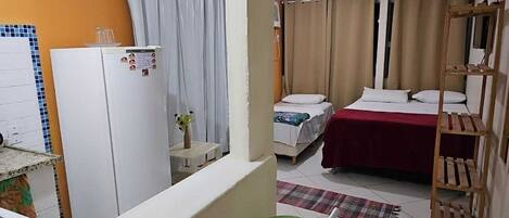 Une chambre d’hôtel compacte comprenant un coin cuisine, un lit recouvert d’une courtepointe rouge, une étagère en bois et une petite chaise verte.
