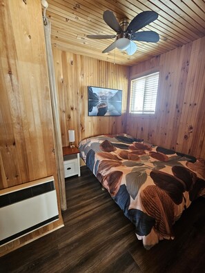 Ein Schlafzimmer einer Holzhütte mit einem Bett, einem Deckenventilator, einem an der Wand montierten Fernseher und einem Fenster.