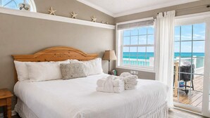 A bedroom with a wooden headboard, white bedding, a view of the ocean through a large window, and a balcony with a chair. - Beachfront Escape | Walk to Trading Post | 4BR (Port St. Joe)