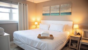 A neatly made bed with white linens, a wooden tray with a rolled towel, two bedside lamps, and a wall with three paintings. - Rehab- Beach front, Sleeps 9 (Port St. Joe)
