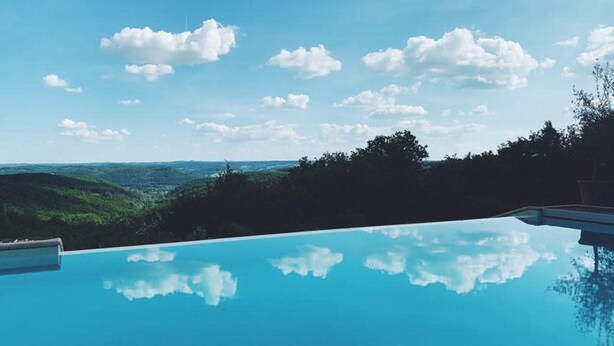 Kirkas, sininen taivas pilvineen, uima-allas, joka heijastaa taivasta, ja näkymä vihreille kukkuloille kauempana.