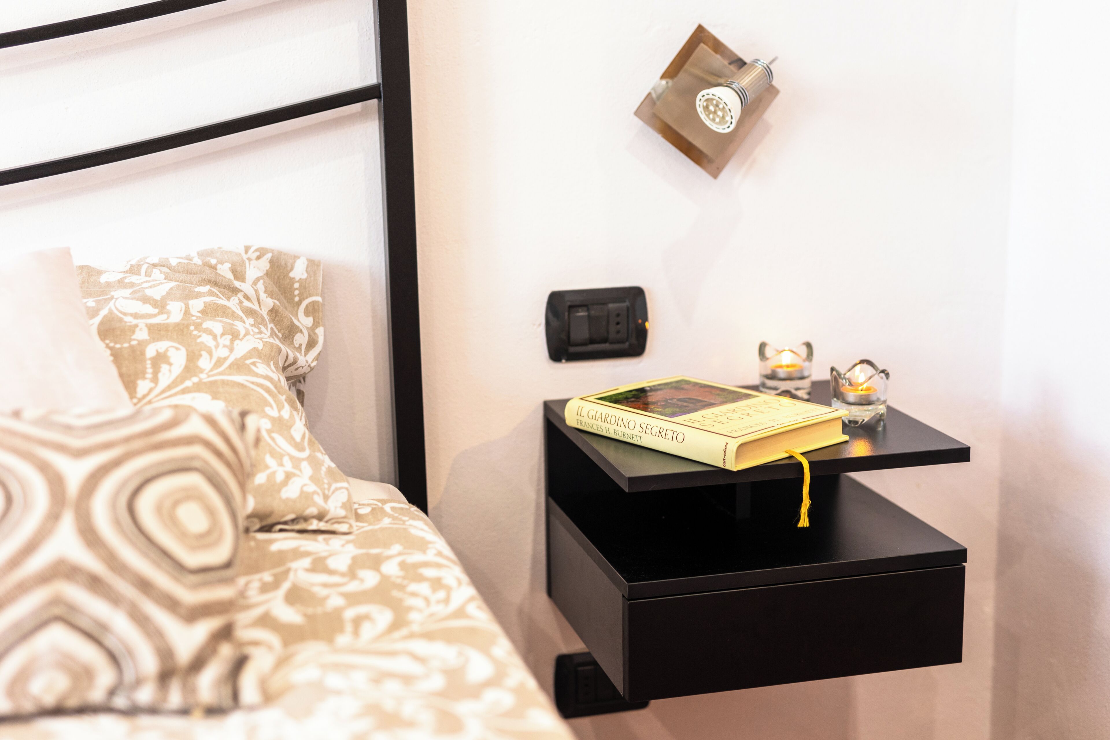 Una cama bien hecha con ropa de cama estampada, una mesita de noche negra con un libro, un enchufe y un pequeño adorno.