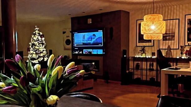 A living room with a television, a Christmas tree, a vase with flowers, and a fruit basket.