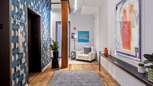 A hallway with a patterned wall, a potted plant, a white chair, a framed artwork, and a wooden floor.