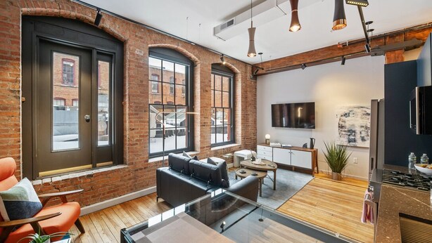 A modern loft with exposed brick walls, large windows, a kitchen area, and a seating area with a glass table.