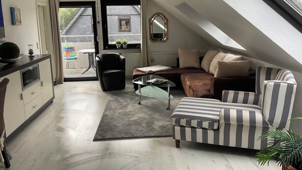 A loft-style bedroom with a sloped ceiling, a sofa, a chair, a small table, and a view of the outside.