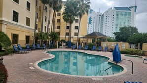 Una piscina con sillas de descanso, una zona de estar techada y un edificio de varios pisos al fondo.