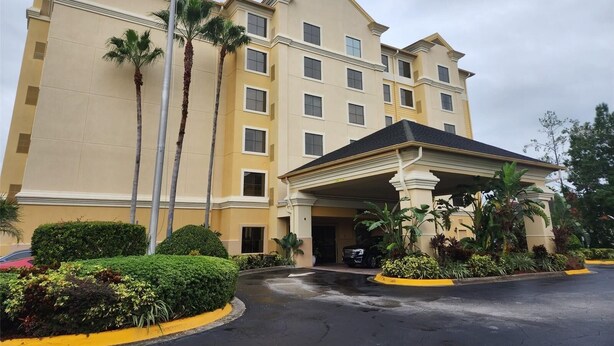 Un hotel de varios pisos con entrada central, palmeras y un área de estacionamiento.