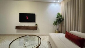 A modern living room with a flat-screen TV mounted on the wall, a glass-top coffee table, a beige sofa with a red cushion, and a potted plant. - Cosy & Chic Apartment with Secure Parking in the Heart of Casablanca (Dar-el-Beida)