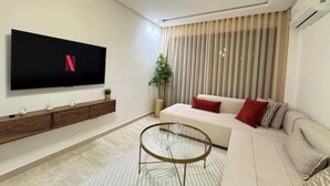 A modern living room with a flat-screen TV, a sofa, a coffee table, and a large window with blinds. - Cosy & Chic Apartment with Secure Parking in the Heart of Casablanca (Dar-el-Beida)