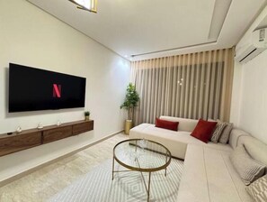 A modern living room with a flat-screen TV, a sofa, a coffee table, and a large window with blinds. - Cosy & Chic Apartment with Secure Parking in the Heart of Casablanca (Dar-el-Beida)
