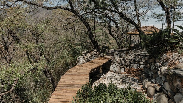 A wooden pathway leading to a stone structure with a thatched roof, surrounded by dense vegetation and hills in the background. - Quinta Real San Carlos (Santiago)