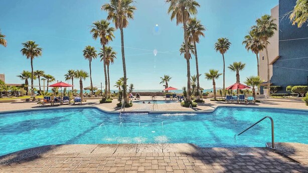 Une piscine bordée de palmiers, avec des parasols et un ciel dégagé.
