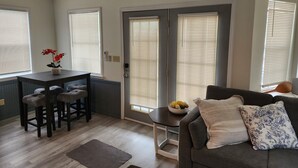 Una sala de estar con un sofá, una mesa de centro con un cuenco de fruta, un comedor con mesa y sillas, y una puerta con ventana.