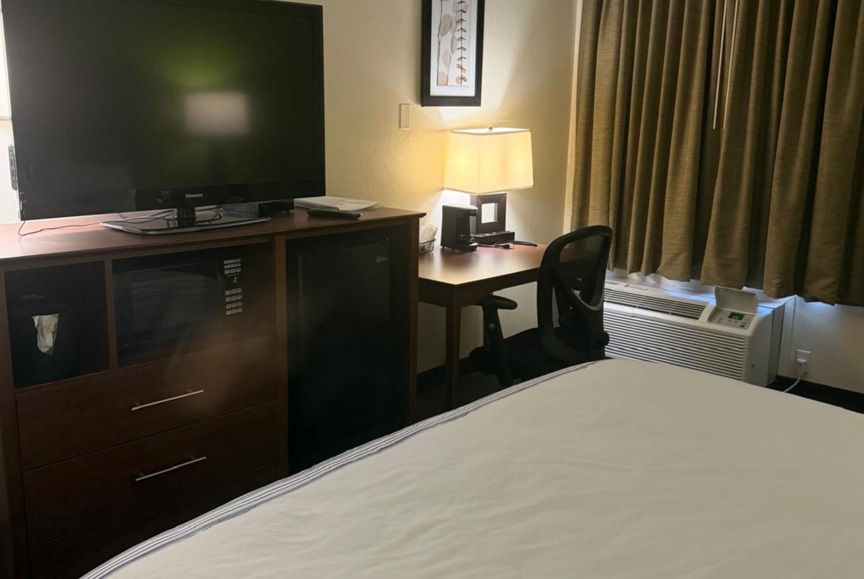 A hotel room with a bed, a desk with a lamp, a television, a wardrobe, and a window with curtains.
