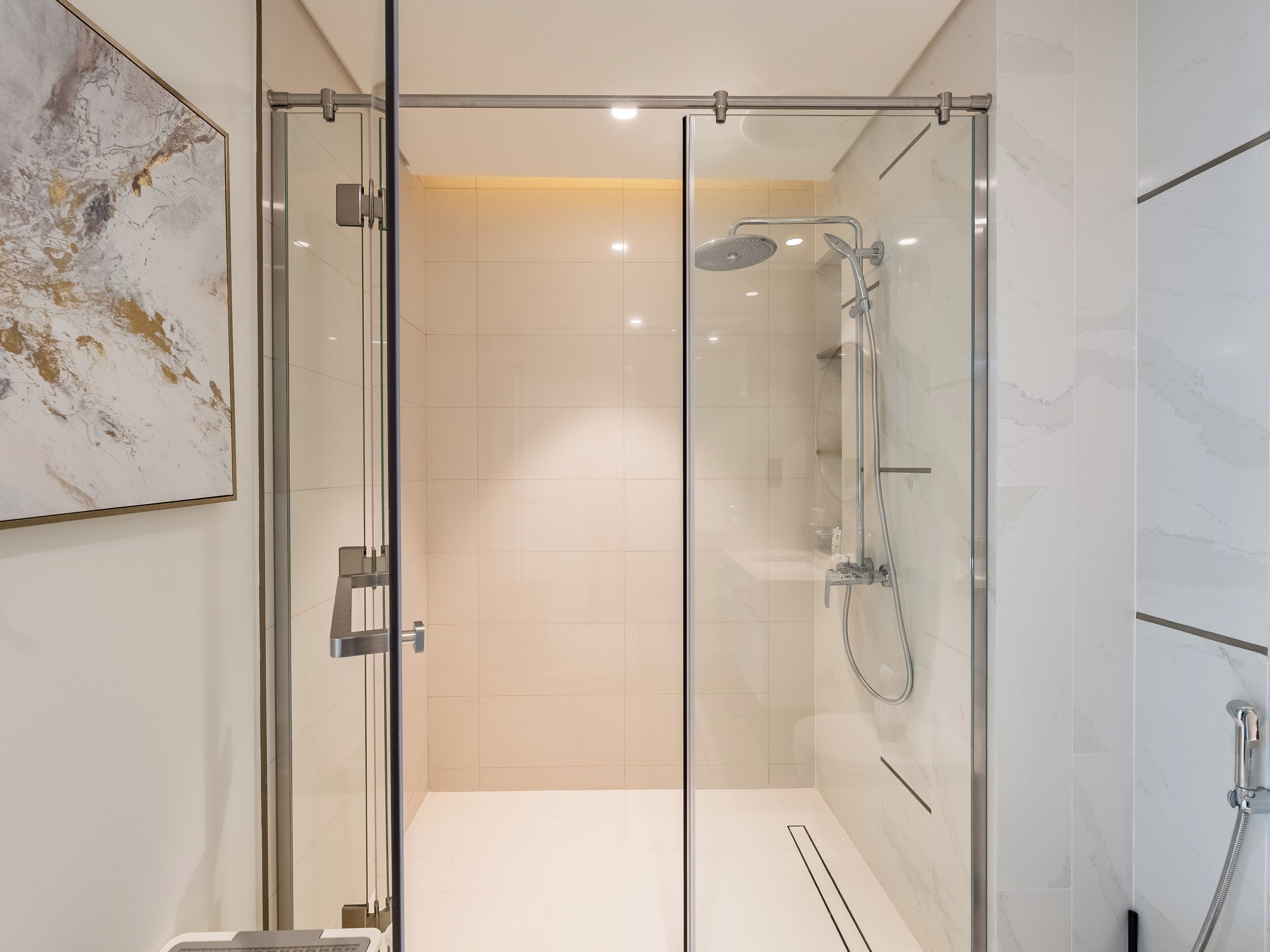 A modern bathroom with a glass-enclosed shower, white marble tiles, and a mounted artwork.