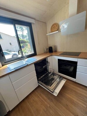 A modern kitchen with a dishwasher, oven, and a window overlooking a residential area. - N&L Signature - Ô Lodge (Mornas)