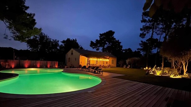 Una piscina con cubierta de madera, una casa con iluminación exterior y árboles al fondo.
