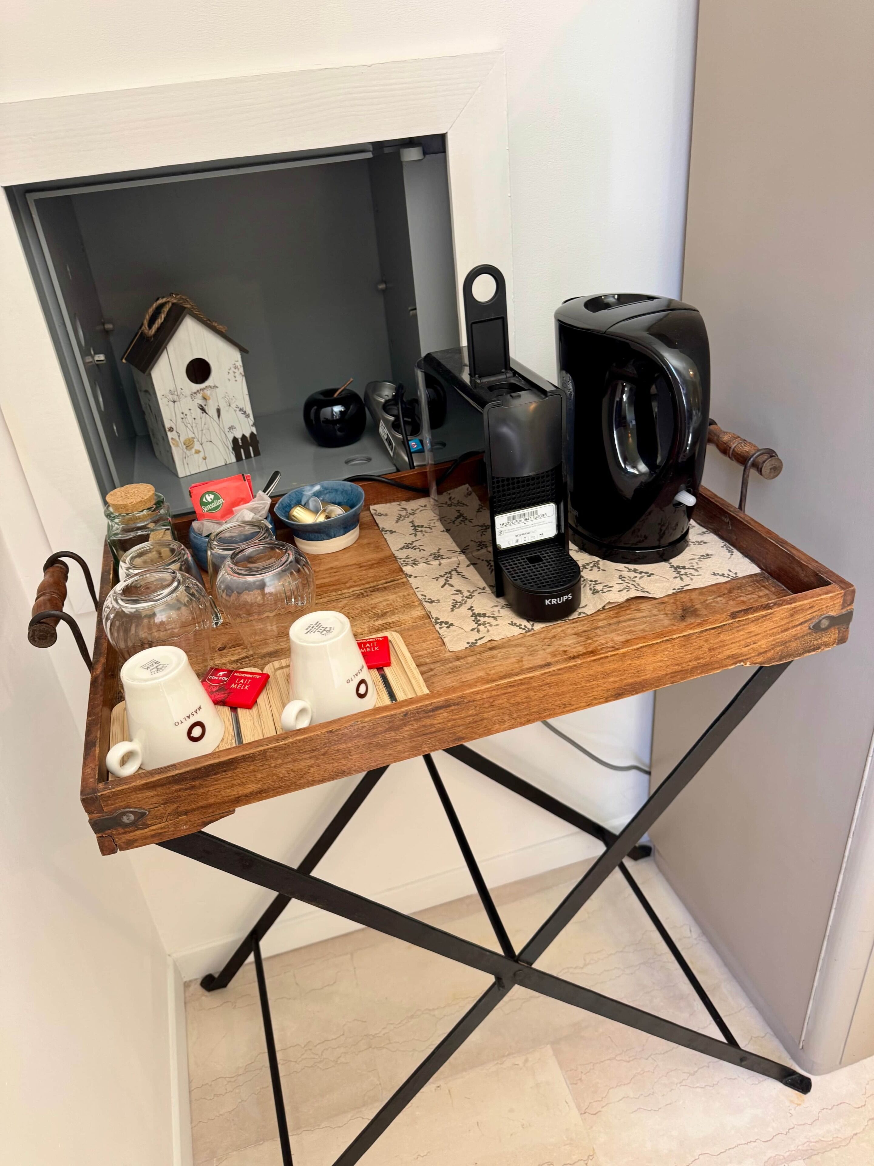 A wooden tray with a coffee maker, jars, and mugs on a metal stand in a room with a white wall and a door.