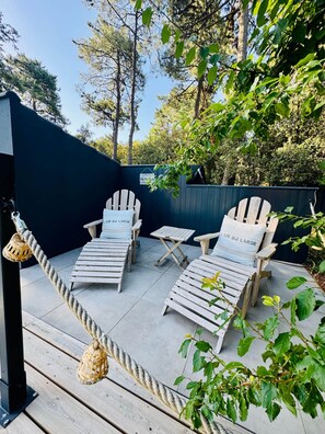 Une terrasse avec deux fauteuils Adirondack portant l’inscription « L’AIR DU LARGE » et une petite table, le tout entouré d’une barrière faite de cordes et d’arbres.