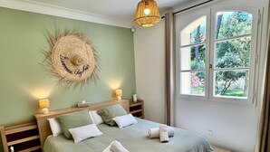 A neatly made bed with pillows and towels, a bedside lamp, a wall-mounted decorative item, and a window with a view of greenery.
