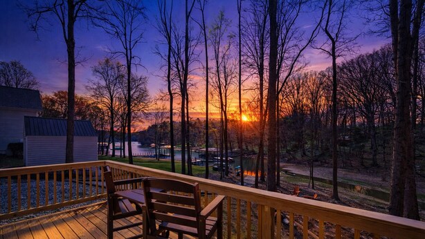 Una terraza de madera con sillas, con vista a un lago y árboles durante el atardecer.
