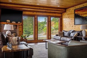 A cozy living room with a fireplace, leather sofas, and large windows offering a view of greenery. - 6-Bedroom Cabin with Indoor Pool, Home Theater & Game Room Near Dollywood (Sevierville)
