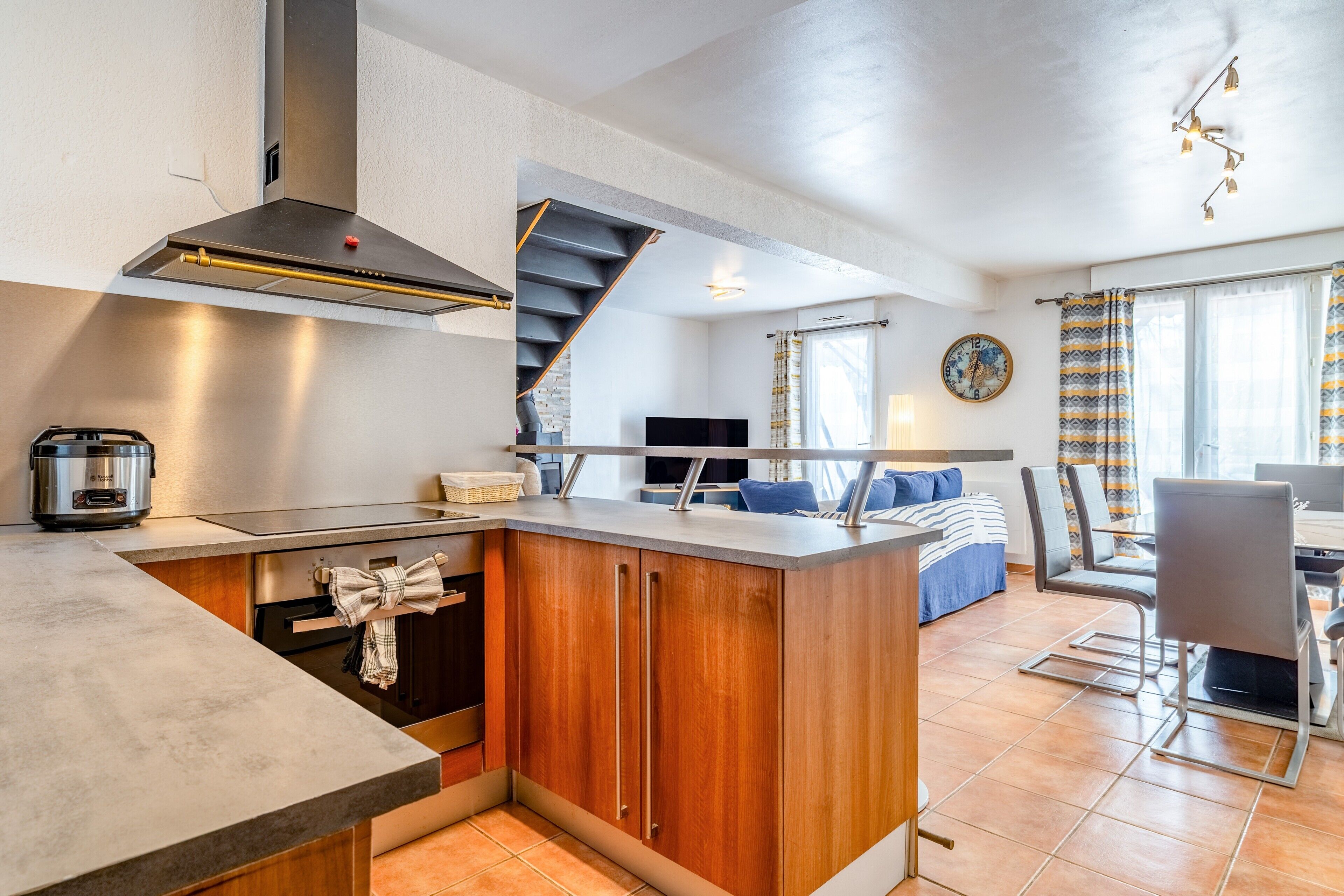 A modern kitchen with a central island, a dining area with a table and chairs, a staircase, and a living room with a sofa and a TV.