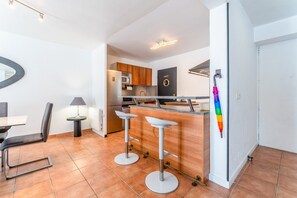 Una cocina moderna con microondas, armarios de madera y un refrigerador negro. Hay dos taburetes blancos y una mesa de comedor con sillas.