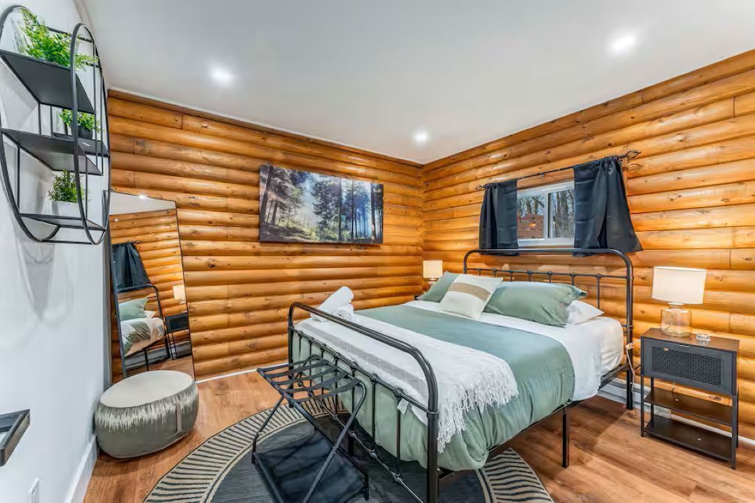 Ein Schlafzimmer in einer Blockhütte mit einem Bett, einem kleinen Tisch, einem Stuhl, einem Spiegel und einem Gemälde an der Wand.
