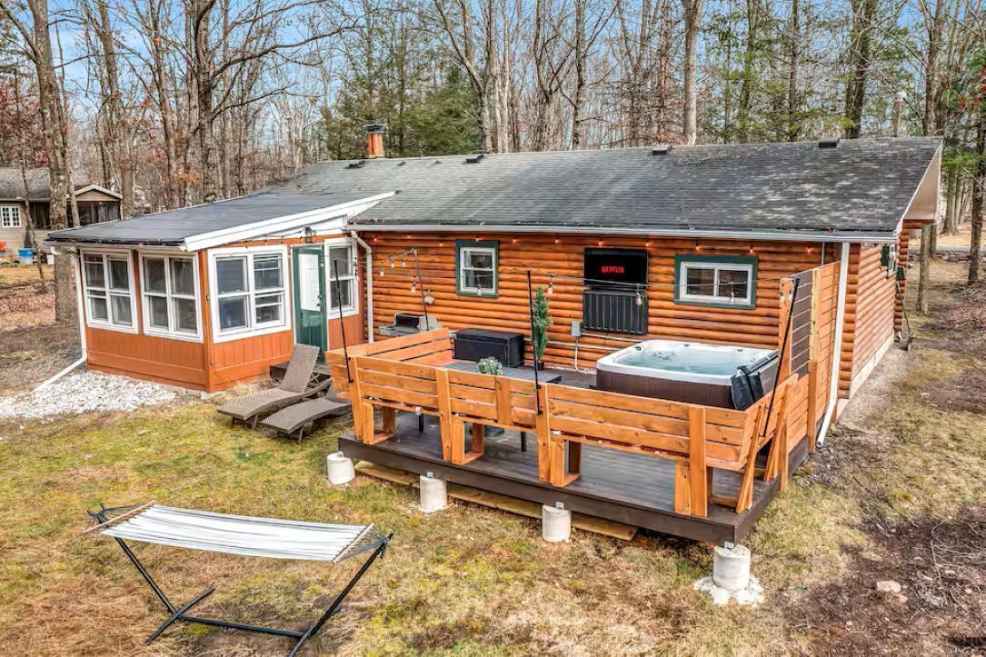 A log cabin with a deck, a hot tub, and a small attached building with windows.