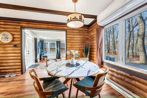A log cabin interior with a round marble table, wooden chairs, a hanging light fixture, and a large window with a forest view. - Log Cabin Retreat W/ Hot Tub in Poconos/Jim Thorpe (Albrightsville)