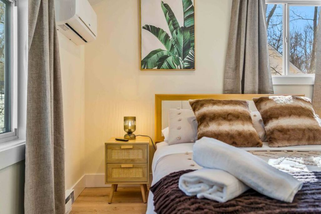 A bedroom with a bed, pillows, a bedside table with a lamp, and a wall-mounted air conditioner.